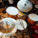 Professional drum set with natural wood finish and red cushioned throne in a studio setting, surrounded by cymbals, percussion gear, and hardwood flooring.